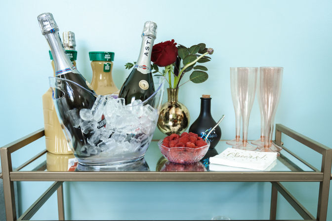 Home bar cart against a teal wall with an ice-filled bucket of sparkling wine, orange juice bottles, a gold vase with red roses, a bowl of raspberries, two champagne flutes and celebratory napkins.