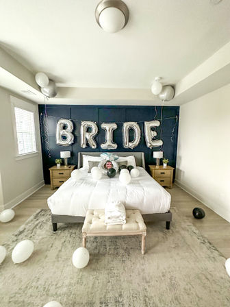Bright master bedroom staged as a bridal suite with silver foil "BRIDE" balloons above crisp white bedding, black-and-white balloons scattered on the floor, wooden nightstands and a tufted bench on a neutral rug.