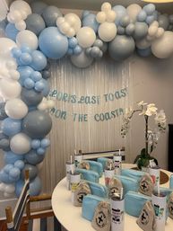Coastal-themed celebration setup with sky-blue, grey and white balloon garland arch, metallic fringe backdrop with teal letter banner, round table of light-blue zip pouches, canvas favor bags, tall tumblers, and a white orchid centerpiece.