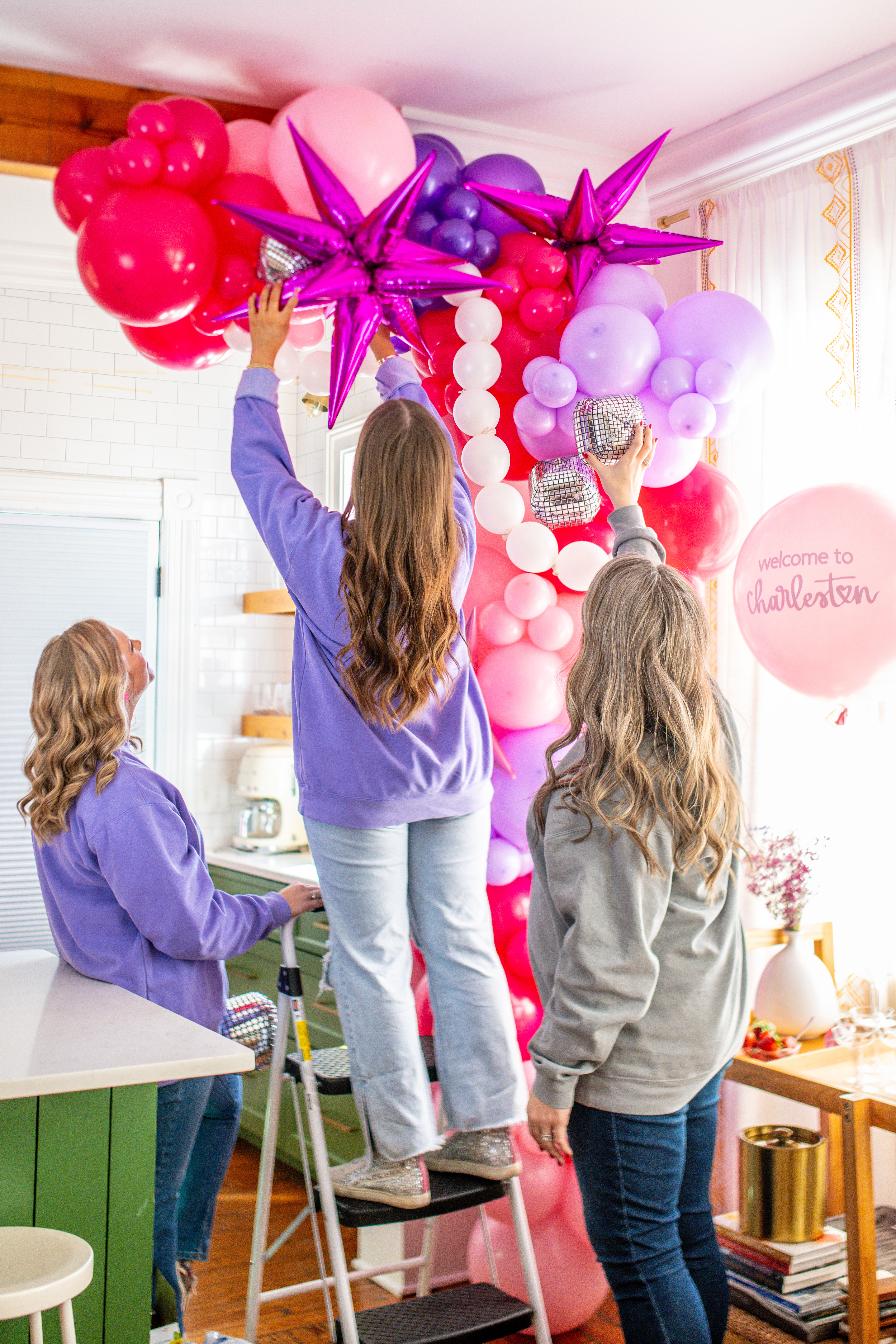 Three people assembling a pink and purple balloon arch with metallic star balloons in a bright Charleston kitchen for party decor