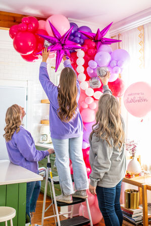Three people assembling a pink and purple balloon arch with metallic star balloons in a bright Charleston kitchen for party decor