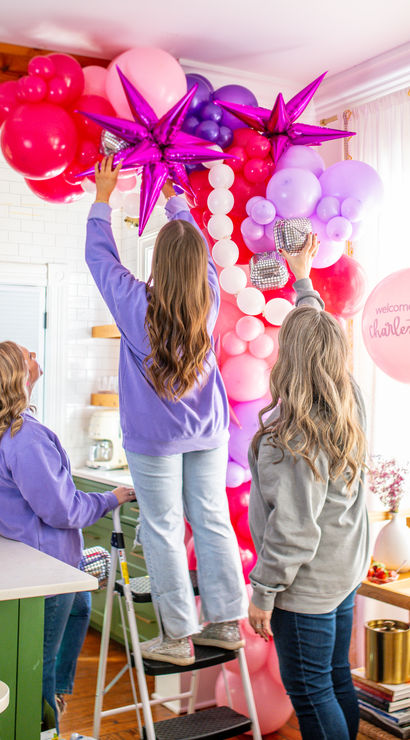 Three people assembling a pink and purple balloon arch with metallic star balloons in a bright Charleston kitchen for party decor