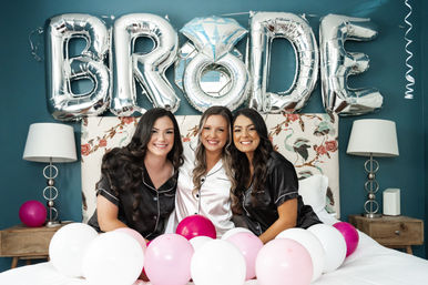 Three women in satin pajamas—bride in white between two bridesmaids in black—smile on a bed in a teal bridal suite with silver "BRIDE" balloons and pink and white balloons