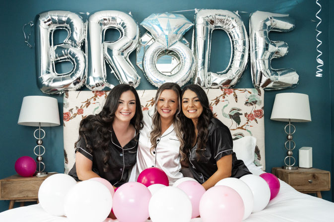 Three women in satin pajamas—bride in white between two bridesmaids in black—smile on a bed in a teal bridal suite with silver "BRIDE" balloons and pink and white balloons