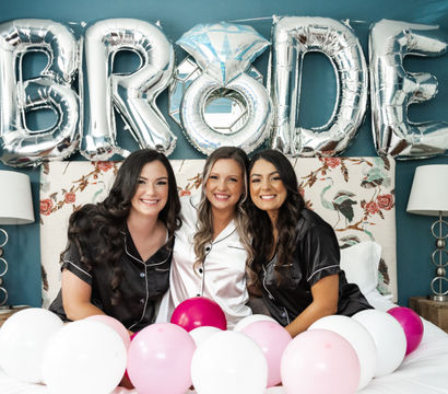 Three women in satin pajamas—bride in white between two bridesmaids in black—smile on a bed in a teal bridal suite with silver "BRIDE" balloons and pink and white balloons