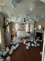 Cozy bridal suite surprise with large white 'BRIDE' letter balloons over a bed and dozens of light blue and pearl balloons floating and scattered across a neutral bedroom with hardwood floors, ceiling fan, and soft natural light.
