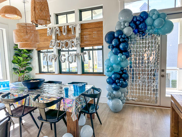 Coastal-style dining room party with a blue balloon arch, silver letter balloons on the windows, rattan pendant lights, holographic table cover and blue cups
