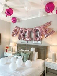 Rose-gold foil "BRIDE" balloons with white and fuchsia balloons floating above a neatly made bed with green pillows and bedside lamps — bridal shower bedroom decor.