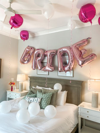 Rose-gold foil "BRIDE" balloons with white and fuchsia balloons floating above a neatly made bed with green pillows and bedside lamps — bridal shower bedroom decor.