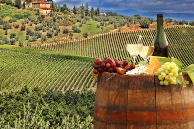 Wooden wine barrel topped with a bottle of white wine, two poured glasses, cheese and red and green grapes, set against rolling vine-covered hills and a farmhouse in a sunlit Tuscan vineyard