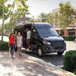 Black Mercedes passenger van parked on a sunny, palm-lined urban shopping street as four casually dressed adults step onto a brick sidewalk from the shuttle near storefronts.