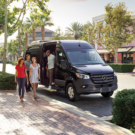 Black Mercedes passenger van parked on a sunny, palm-lined urban shopping street as four casually dressed adults step onto a brick sidewalk from the shuttle near storefronts.