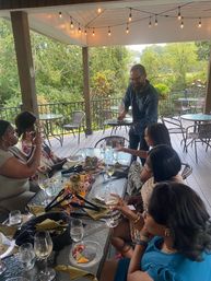 Adults enjoying wine and charcuterie on a covered outdoor patio with string lights and leafy green view as a server places a glass — casual al fresco dining and wine tasting scene.