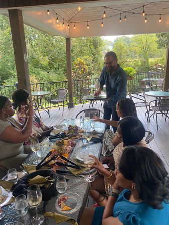 Adults enjoying wine and charcuterie on a covered outdoor patio with string lights and leafy green view as a server places a glass — casual al fresco dining and wine tasting scene.