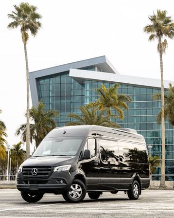 Sleek black passenger van parked in front of a modern glass building framed by tall palm trees in a sunny coastal cityscape