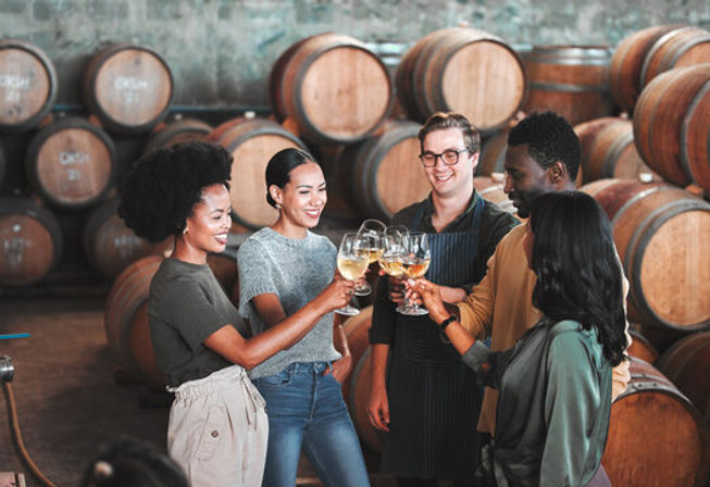 Group of five friends smiling and clinking glasses of white wine during a cheerful wine tasting in a barrel-lined winery cellar