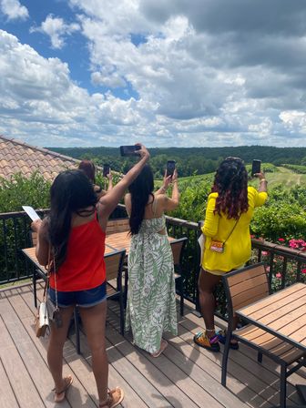 Group of women on a sunny vineyard deck snapping photos of rolling green hills and a dramatic cloudy blue sky