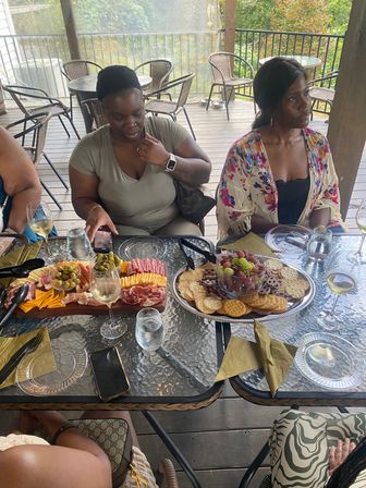 Outdoor patio scene with two people at a glass-top table on a wooden deck enjoying charcuterie boards of cheese, cured meats, olives, grapes and crackers, with wine glasses and wicker chairs in the background.