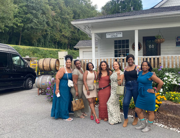Seven women smiling and posing outside a white winery tasting room on a porch with stacked wooden barrels, colorful flower beds, and a black van nearby — casual summer outfits, group photo.