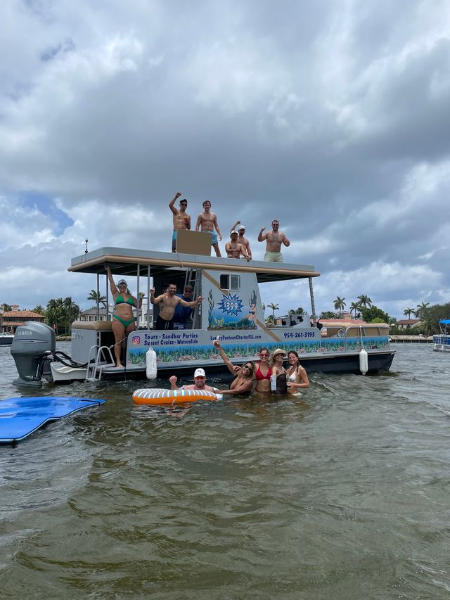 Group of friends having a lively boat party on a double-decker pontoon in a Florida coastal inlet, some on the roof and others in the water on floats under a cloudy sky with waterfront homes and palm trees in the background.