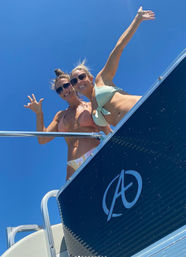 Two smiling women in bikinis waving from the deck of a boat under a clear blue sky on a sunny summer boating trip.