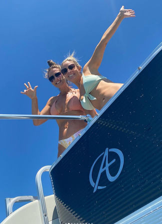 Two smiling women in bikinis waving from the deck of a boat under a clear blue sky on a sunny summer boating trip.