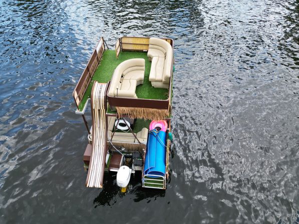 Aerial view of a compact pontoon boat on rippled water with green turf deck, curved beige lounge seating, a waterslide, blue rolled float and pink inner tube, plus an outboard motor.