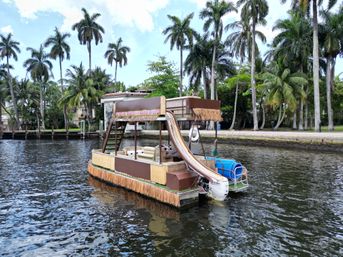 Tiki-style pontoon boat with waterslide and thatched trim floating on a palm‑lined Florida waterfront canal with coastal homes in the background.