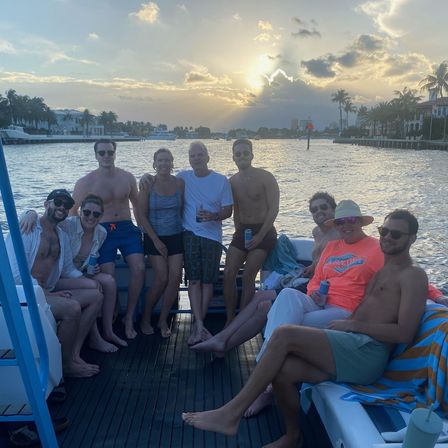 Nine people relaxing on a boat during a palm-lined coastal canal sunset, holding drinks and smiling.