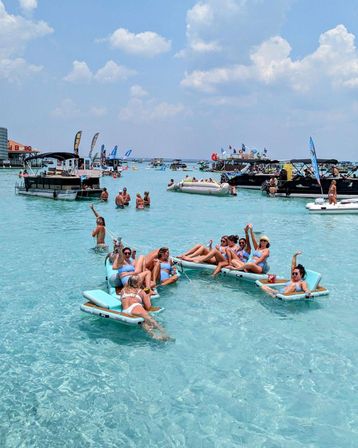 Group of people lounging on connected inflatable floats in shallow clear turquoise water surrounded by anchored party boats and flags under a sunny, cloudy sky — lively summer boat-party beach scene.