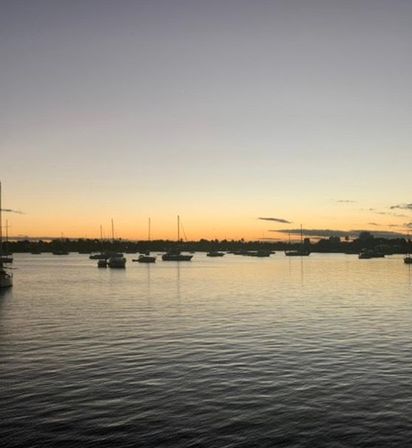 Sunset over a calm coastal harbor with silhouetted sailboats and masts on a golden-orange horizon reflecting on rippling water