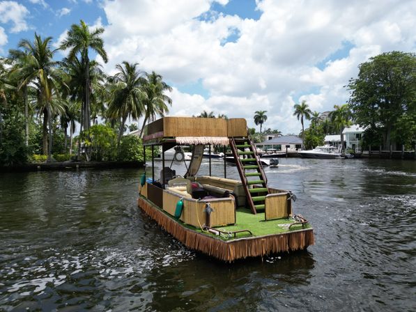 Tiki-style double-decker pontoon boat with grass carpeting and fringe skirts cruising a tropical waterfront canal lined with palm trees, docks, and houses under a blue sky.