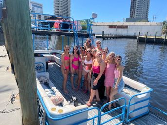 Cheerful group of women in pink swimsuits and summer outfits wearing heart-shaped sunglasses posing on a pontoon boat at a sunny urban marina, docked beside a wooden pier with high-rise buildings in the background.