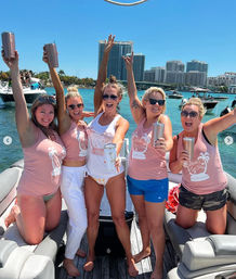 Five friends in matching pink tank tops cheering with tumblers on a boat party in a sunny turquoise bay, with other boats and a coastal high‑rise skyline in the background.