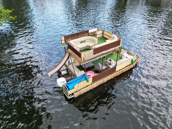 Double-decker tiki-style pontoon party boat with a water slide, turf deck, cushioned lounge seating and inflatable tube floating on a calm river