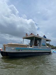 Pontoon party boat with people lounging on the upper deck, colorful sea-themed mural along the side, floating in calm bay water under a dramatic cloudy sky