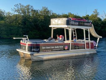 Double-decker pontoon boat with waterslide and two passengers cruising a mangrove-lined coastal Florida waterway in golden-hour light, relaxed sunset cruise scene.
