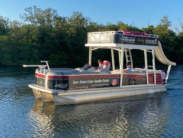 Double-decker pontoon boat with waterslide and two passengers cruising a mangrove-lined coastal Florida waterway in golden-hour light, relaxed sunset cruise scene.