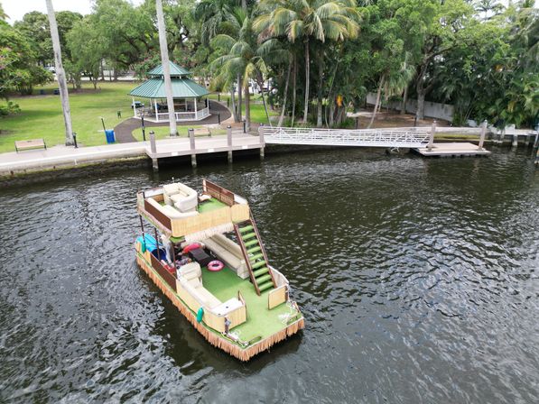 Two-level tiki-themed party boat with green artificial turf, lounge seating and stairs floating on a river beside a palm-lined waterfront park, gazebo and dock.