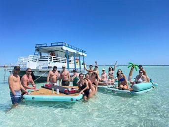 Group of friends partying on inflatable platforms and a floating table in crystal-clear shallow turquoise water beside a two-deck party boat under a bright blue sky – sun, drinks, and sandbar vibes.