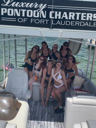 Group of women on a Fort Lauderdale pontoon boat smiling in swimsuits for a bachelorette party, bride-to-be sash, holding cold drinks with American flag and turquoise water in the background.