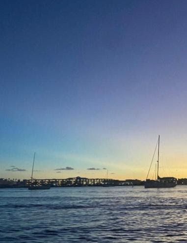 Dusk coastal harbor at sunset with anchored sailboats, calm rippling water, and a lit marina on the horizon
