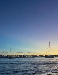 Dusk coastal harbor at sunset with anchored sailboats, calm rippling water, and a lit marina on the horizon