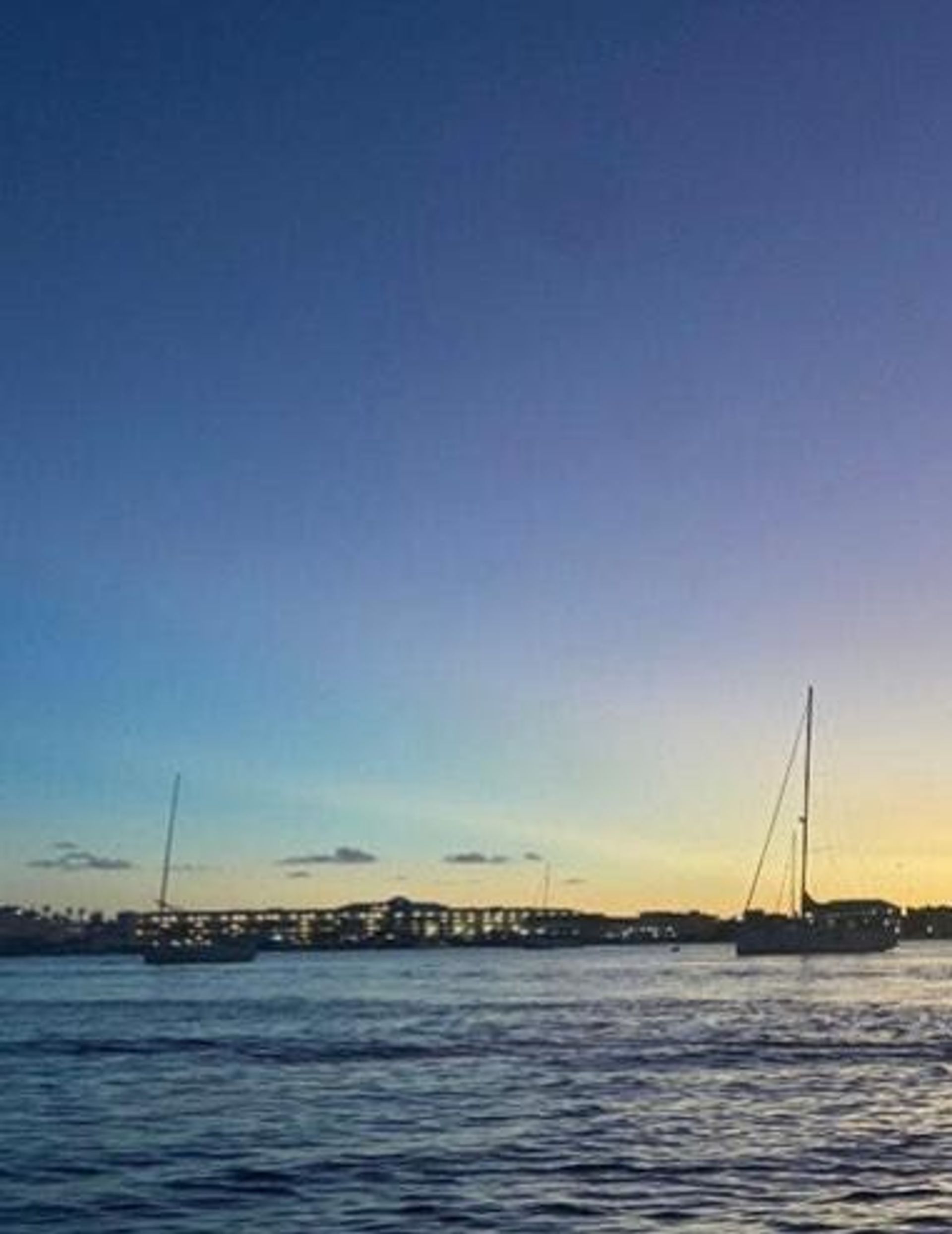 Dusk coastal harbor at sunset with anchored sailboats, calm rippling water, and a lit marina on the horizon