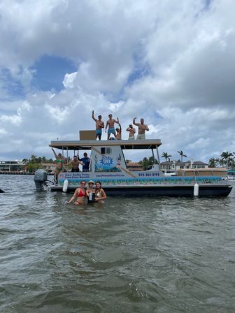 Group of friends cheering on a two-level pontoon boat while others wade in shallow coastal Florida waters, palm trees and waterfront homes under a dramatic cloudy sky