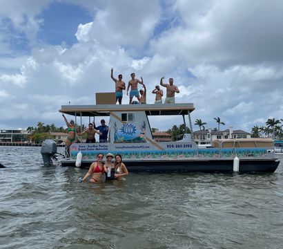 Group of friends cheering on a two-level pontoon boat while others wade in shallow coastal Florida waters, palm trees and waterfront homes under a dramatic cloudy sky