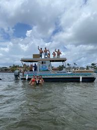 Friends partying on a double-decker pontoon boat and wading in shallow coastal water near palm-lined waterfront homes under a dramatic cloudy sky.