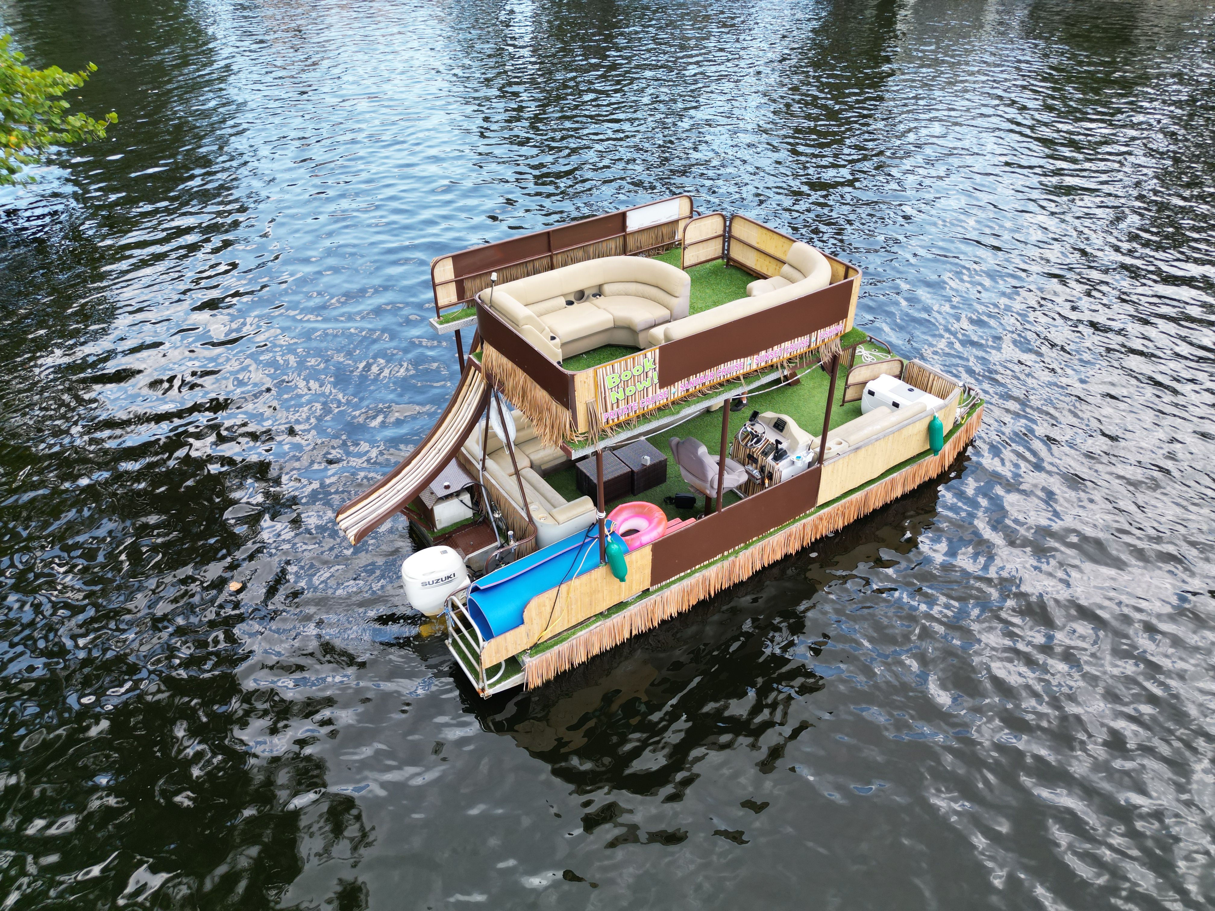 Party-ready double-decker pontoon boat with curved lounge seating, waterslide, tiki fringe and outboard motor floating on a calm river