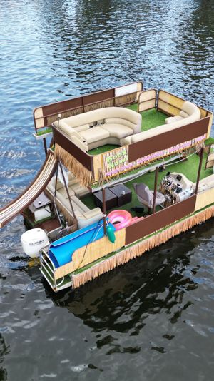 Party-ready double-decker pontoon boat with curved lounge seating, waterslide, tiki fringe and outboard motor floating on a calm river