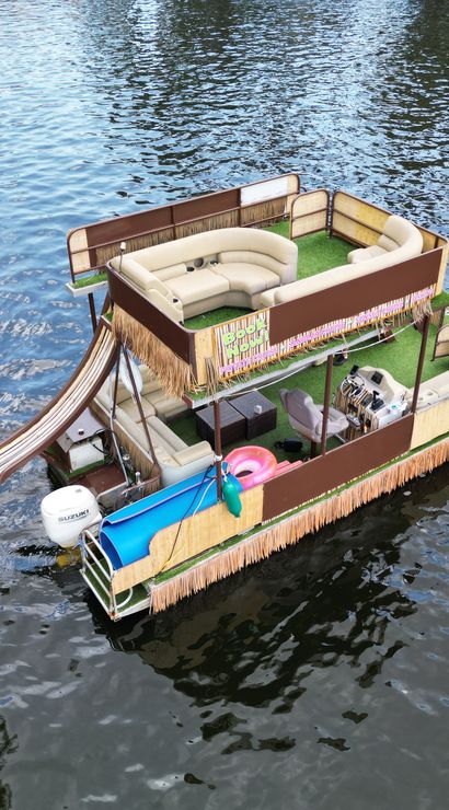 Party-ready double-decker pontoon boat with curved lounge seating, waterslide, tiki fringe and outboard motor floating on a calm river